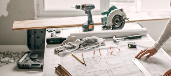 Construction workspace with blueprints, safety gloves, pencils, and tools including a drill, circular saw, and tape measure.